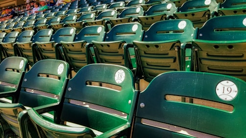 A close-up view of several rows of green stadium seats, each labeled with sequential numbers. The seats are empty, and in the background, a few people can be seen sitting further back.