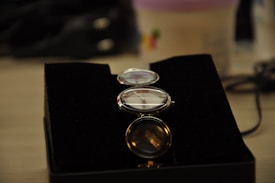 A watch with a white face and silver casing is placed on a black velvet backdrop. The watch features visible clock hands with a red second hand, and small black hour indices. Several additional lenses or magnifiers are placed above it, all with metallic rims.
