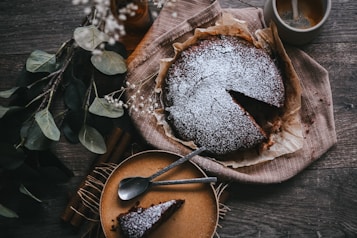 A chocolate cake sprinkled with powdered sugar is placed on a piece of parchment paper. Beside the cake, a slice has been cut and is resting on a small plate with two silver spoons. On the right, a cup of coffee sits with foamy bubbles on top, and on the left, there are eucalyptus leaves and white flowers scattered around, creating a rustic ambiance.