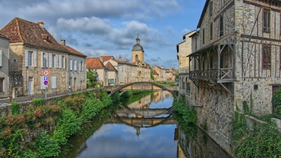A charming European village with historic stone buildings lining a narrow, reflective river. A small bridge arches over the water, leading to a distant church with a prominent dome. Greenery intersperses the scene, adding natural beauty to the architectural view.
