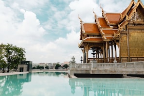A traditional Thai pavilion with intricate wooden carvings and red-tiled roofs stands beside a serene reflecting pool. The sky is filled with fluffy white clouds, providing a picturesque backdrop. Lush green trees are visible in the distance, along with some urban buildings across the water.