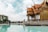 A traditional Thai pavilion with intricate wooden carvings and red-tiled roofs stands beside a serene reflecting pool. The sky is filled with fluffy white clouds, providing a picturesque backdrop. Lush green trees are visible in the distance, along with some urban buildings across the water.