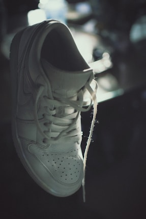 A white sneaker with visible laces is suspended in mid-air, casting subtle shadows under soft lighting. The background is blurred, giving the scene a somewhat moody and artistic impression.