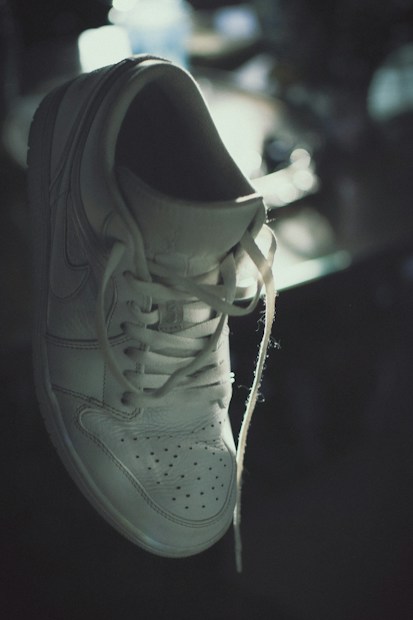 A white sneaker with visible laces is suspended in mid-air, casting subtle shadows under soft lighting. The background is blurred, giving the scene a somewhat moody and artistic impression.