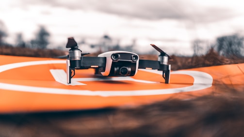 A small drone with propellers is positioned on an orange landing pad outdoors. The background is blurred, indicating an open field with a cloudy sky.