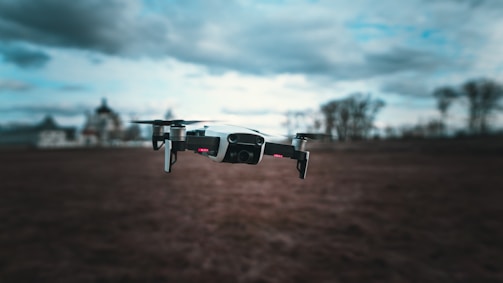 A drone hovers over an open field, with a slightly blurred background featuring distant trees and buildings under a cloudy sky.