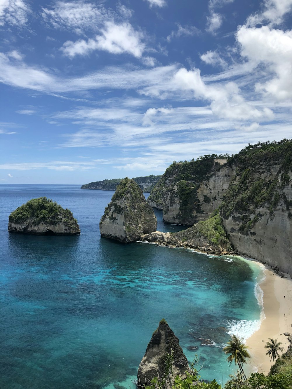 昼間の青と白の曇り空の下、青い海のそばに緑と茶色の岩層