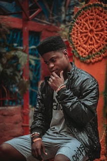 A person is sitting thoughtfully against a colorful, artistic background. The individual is wearing a shiny black jacket and gray shorts with a logo, accessorized with bracelets. The setting includes vibrant red, blue, orange, and green tones, with an intricate circular design on the wall.