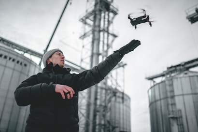 A person wearing a black jacket and beanie is controlling a drone with one hand raised towards the drone. The backdrop includes industrial structures, such as large silos and metal frameworks, suggesting a setting in an industrial or agricultural area.
