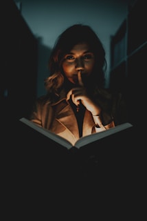 A woman with wavy hair is standing in dim lighting, holding an open book. The light from the book illuminates her face partially, creating a mysterious atmosphere. Her finger is raised to her lips, suggesting silence or secrecy.