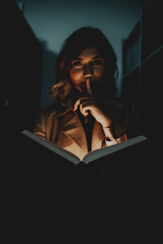 A woman with wavy hair is standing in dim lighting, holding an open book. The light from the book illuminates her face partially, creating a mysterious atmosphere. Her finger is raised to her lips, suggesting silence or secrecy.