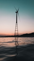 A wind turbine is silhouetted against a gradient sky that transitions from light peach near the horizon to a deeper teal above. The turbine stands on a metal tower, rising from a body of still water reflecting the muted colors of the sky.