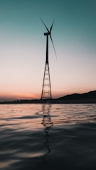 A wind turbine is silhouetted against a gradient sky that transitions from light peach near the horizon to a deeper teal above. The turbine stands on a metal tower, rising from a body of still water reflecting the muted colors of the sky.