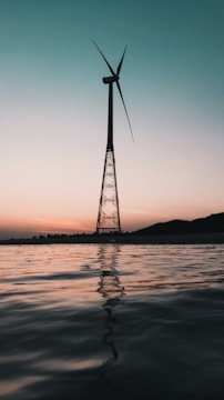 A wind turbine is silhouetted against a gradient sky that transitions from light peach near the horizon to a deeper teal above. The turbine stands on a metal tower, rising from a body of still water reflecting the muted colors of the sky.