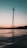 A wind turbine is silhouetted against a gradient sky that transitions from light peach near the horizon to a deeper teal above. The turbine stands on a metal tower, rising from a body of still water reflecting the muted colors of the sky.