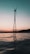 A wind turbine is silhouetted against a gradient sky that transitions from light peach near the horizon to a deeper teal above. The turbine stands on a metal tower, rising from a body of still water reflecting the muted colors of the sky.