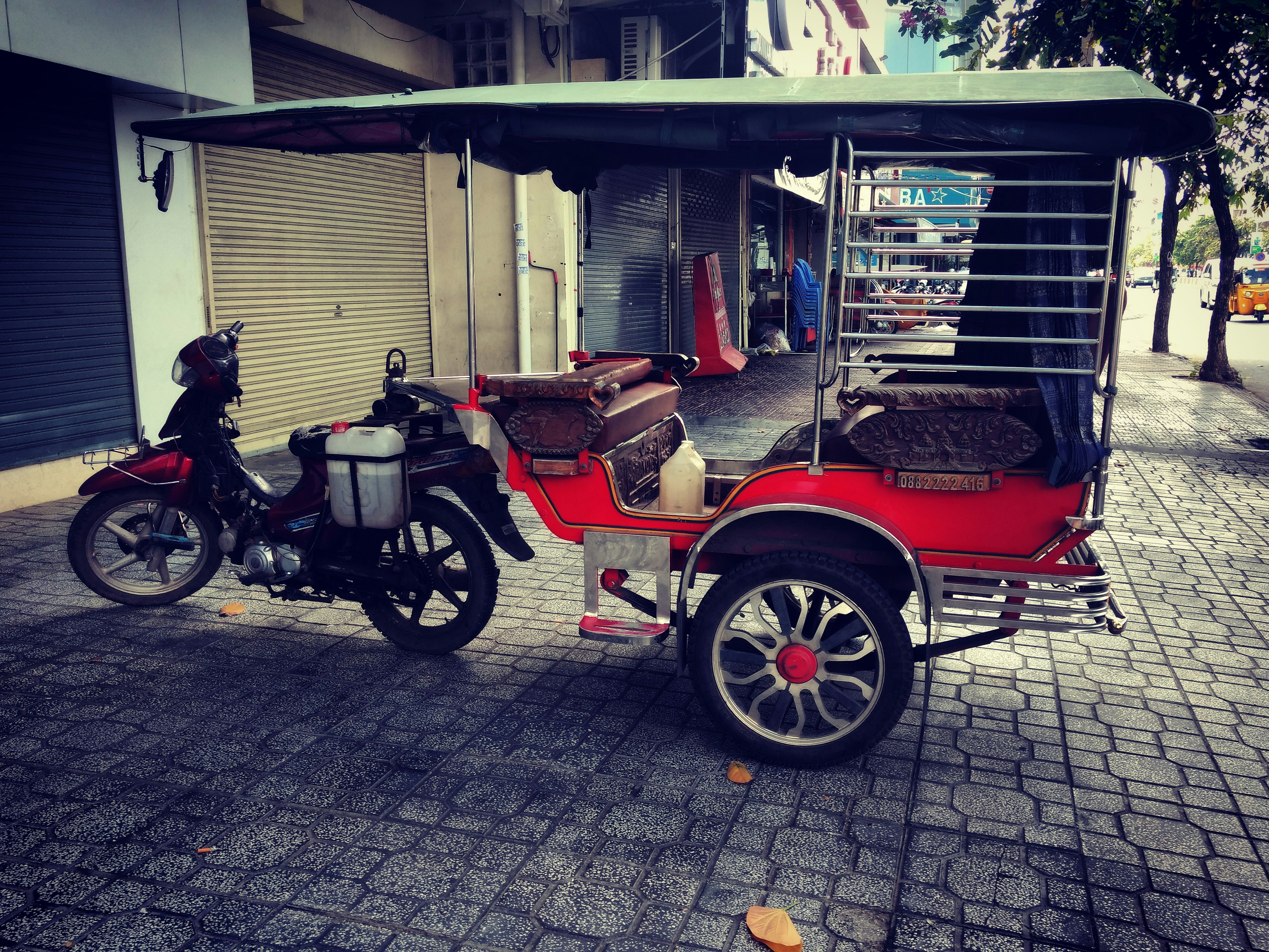 A motorcycle attached to a vibrant red rickshaw parked on a cobblestone sidewalk. The rickshaw has a canopy and cushioned seats, with chrome details and large spoked wheels. The background features closed shop shutters and some scattered leaves on the ground.