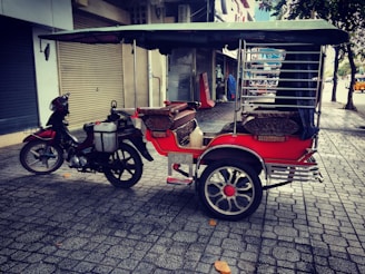 A motorcycle attached to a vibrant red rickshaw parked on a cobblestone sidewalk. The rickshaw has a canopy and cushioned seats, with chrome details and large spoked wheels. The background features closed shop shutters and some scattered leaves on the ground.