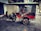 A motorcycle attached to a vibrant red rickshaw parked on a cobblestone sidewalk. The rickshaw has a canopy and cushioned seats, with chrome details and large spoked wheels. The background features closed shop shutters and some scattered leaves on the ground.
