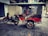 A motorcycle attached to a vibrant red rickshaw parked on a cobblestone sidewalk. The rickshaw has a canopy and cushioned seats, with chrome details and large spoked wheels. The background features closed shop shutters and some scattered leaves on the ground.