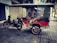 A motorcycle attached to a vibrant red rickshaw parked on a cobblestone sidewalk. The rickshaw has a canopy and cushioned seats, with chrome details and large spoked wheels. The background features closed shop shutters and some scattered leaves on the ground.