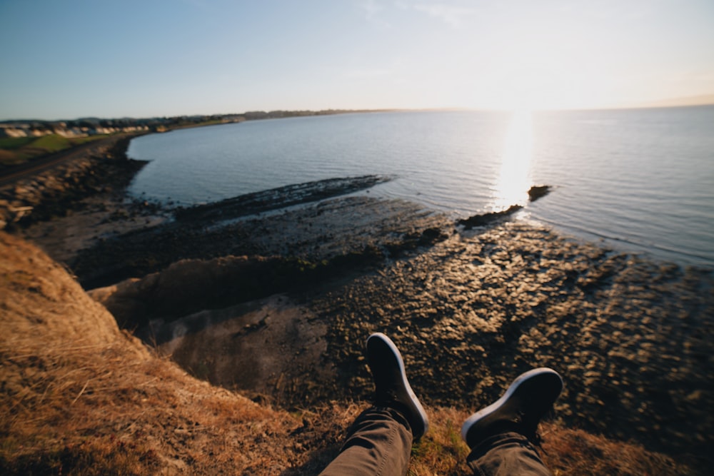 Person in schwarzen Hosen und schwarzen Schuhen sitzt auf braunem Felsen in der Nähe von Gewässern während