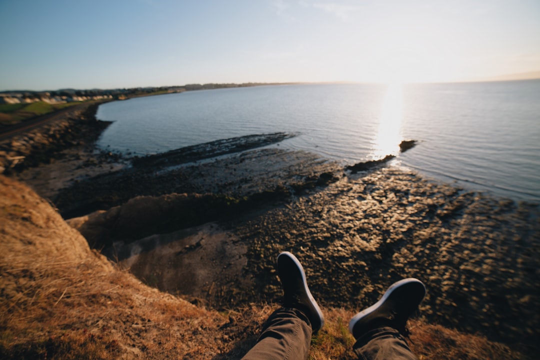 person in black pants and black shoes sitting on brown rock near body of water during