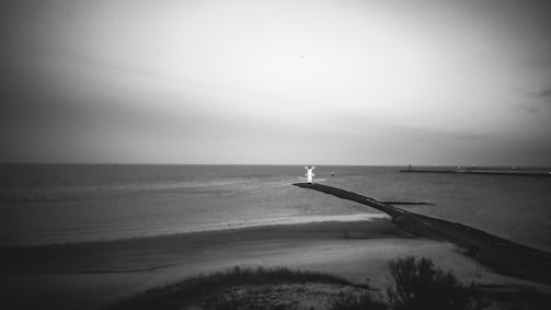 A monochrome image of a coastal landscape with a prominent statue standing at the edge of a long pier extending into the sea. The shoreline features sandy beaches and some sparse vegetation in the foreground. The sky is overcast, with the horizon line visible where the sea meets the sky.