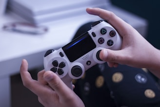 A person is holding a white game controller, specifically a PlayStation controller, in both hands. The background features a blurred white entertainment system or console. The person is wearing dark pants with patterns of circles and lines.
