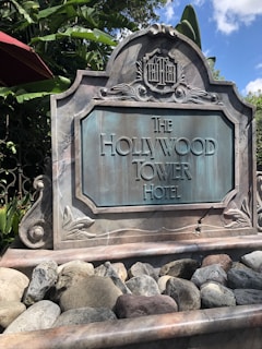 A sign for The Hollywood Tower Hotel crafted in a vintage style with decorative elements, set against a background of green foliage and clear skies.