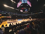 A basketball game is taking place in a large arena filled with spectators. The players are on the court, with one team wearing yellow jerseys. Overhead, a large digital scoreboard displays scores and advertisements. The audience is seated close to the court, watching intently.