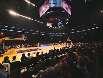 A basketball game is taking place in a large arena filled with spectators. The players are on the court, with one team wearing yellow jerseys. Overhead, a large digital scoreboard displays scores and advertisements. The audience is seated close to the court, watching intently.