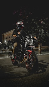 A motorcyclist wearing a helmet sits on a sleek, modern motorbike parked on a dimly lit street at night. The bike is black with red accents, and the headlight is on, illuminating the immediate area. A large building with lit windows is visible in the background. The scene conveys a sense of adventure and mystery.