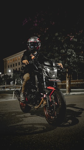 A motorcyclist wearing a helmet sits on a sleek, modern motorbike parked on a dimly lit street at night. The bike is black with red accents, and the headlight is on, illuminating the immediate area. A large building with lit windows is visible in the background. The scene conveys a sense of adventure and mystery.