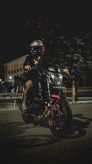 A motorcyclist wearing a helmet sits on a sleek, modern motorbike parked on a dimly lit street at night. The bike is black with red accents, and the headlight is on, illuminating the immediate area. A large building with lit windows is visible in the background. The scene conveys a sense of adventure and mystery.