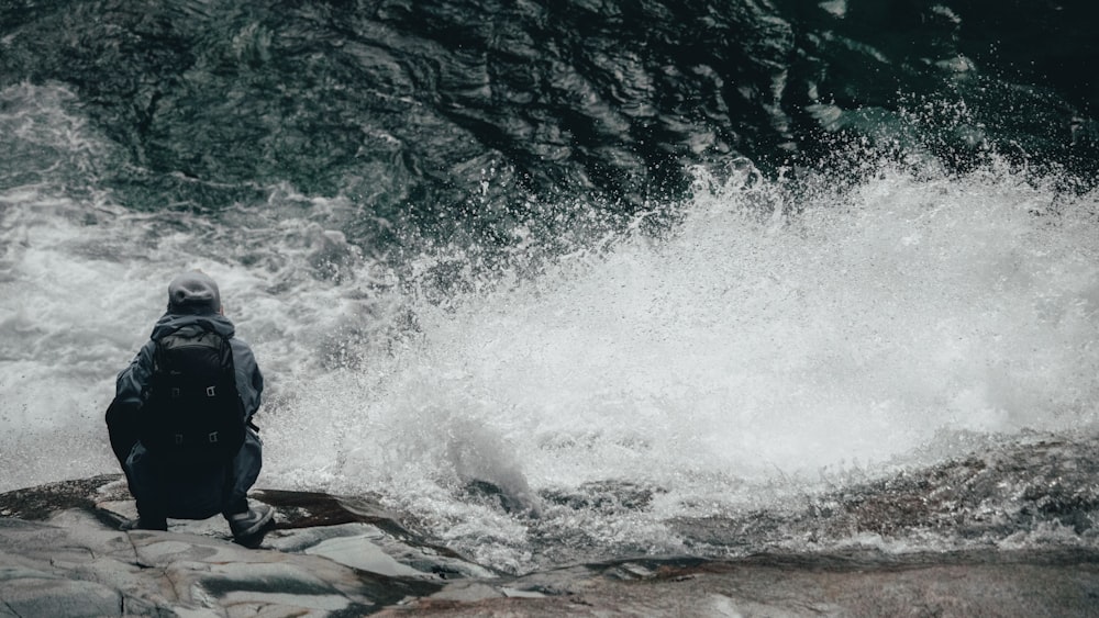 man in black jacket sitting on rock near water waves during daytime