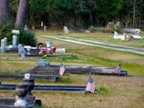 A serene graveyard setting with tombstones and memorial vases lined along grassy pathways, adorned with small American flags. Some graves are decorated with bouquets of flowers and assorted ornamental objects. In the background, trees form a dense green barrier.