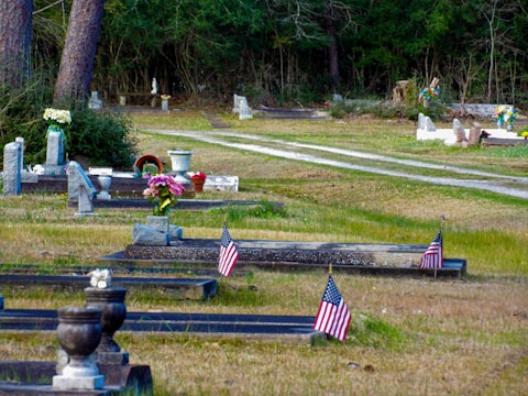 A serene graveyard setting with tombstones and memorial vases lined along grassy pathways, adorned with small American flags. Some graves are decorated with bouquets of flowers and assorted ornamental objects. In the background, trees form a dense green barrier.
