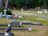 A serene graveyard setting with tombstones and memorial vases lined along grassy pathways, adorned with small American flags. Some graves are decorated with bouquets of flowers and assorted ornamental objects. In the background, trees form a dense green barrier.