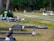 A serene graveyard setting with tombstones and memorial vases lined along grassy pathways, adorned with small American flags. Some graves are decorated with bouquets of flowers and assorted ornamental objects. In the background, trees form a dense green barrier.
