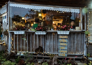 A cozy café setting with a rustic wooden exterior, adorned with string lights and vibrant flowers in planters. Inside, two people engage in activities with warm lighting. A handwritten menu sign hangs on the outside.
