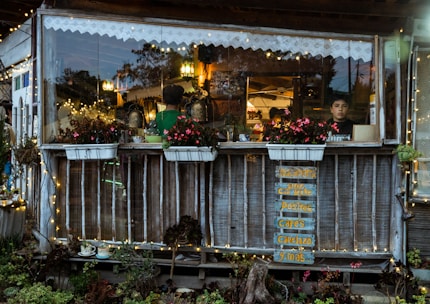 A cozy café setting with a rustic wooden exterior, adorned with string lights and vibrant flowers in planters. Inside, two people engage in activities with warm lighting. A handwritten menu sign hangs on the outside.
