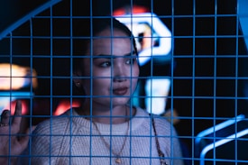 A person with a thoughtful expression stands behind a blue wire grid. The background features blurred neon lights and signage, creating a moody atmosphere. The individual is wearing a light-colored sweater and gold jewelry, touching the grid gently.