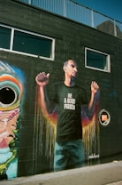 A mural painted on a black brick wall features a person raising their fists and wearing a shirt with the words 'Be A Good Person'. The mural includes vibrant colors dripping downwards, creating a dynamic and lively effect. Above the mural are windows and a fence, suggesting it's part of an urban building.