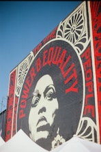 A large mural on a brick wall features a striking black-and-white portrait of a woman surrounded by bold red typography. The words 'Power & Equality' and 'Power to the People' are prominently displayed. Decorative patterns frame the portrait, adding artistic flair to the composition.