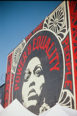A large mural on a brick wall features a striking black-and-white portrait of a woman surrounded by bold red typography. The words 'Power & Equality' and 'Power to the People' are prominently displayed. Decorative patterns frame the portrait, adding artistic flair to the composition.
