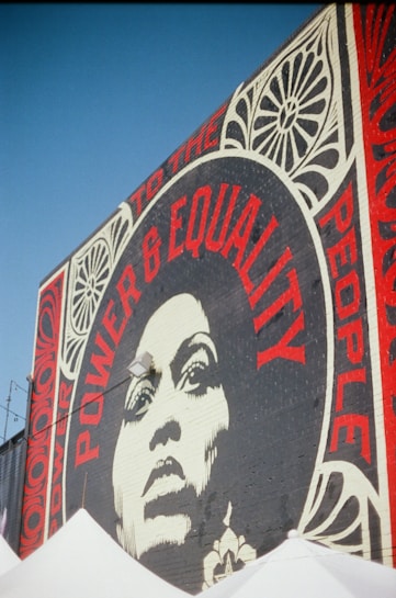 A large mural on a brick wall features a striking black-and-white portrait of a woman surrounded by bold red typography. The words 'Power & Equality' and 'Power to the People' are prominently displayed. Decorative patterns frame the portrait, adding artistic flair to the composition.