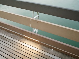 A wooden boardwalk with parallel planks leads to a metal railing. The railing consists of horizontal beams painted in beige and a darker top rail, with water visible behind it in a muted green shade. Sunlight creates a soft reflection on the surface, adding warmth to the scene.