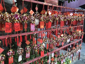 A display of colorful charms and ornaments is hanging on a red wooden frame. The ornaments are spherical, decorated with golden and pink elements, and accompanied by red tassels and printed tags with Asian characters.