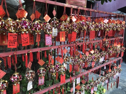 A display of colorful charms and ornaments is hanging on a red wooden frame. The ornaments are spherical, decorated with golden and pink elements, and accompanied by red tassels and printed tags with Asian characters.